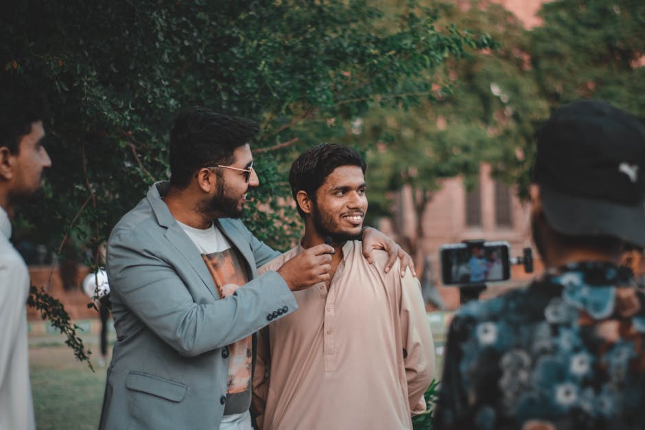 Two men smiling with one adjusting the other's collar as a third person captures the moment on a smartphone camera. They are outdoors, surrounded by trees, with a brick building in the background.