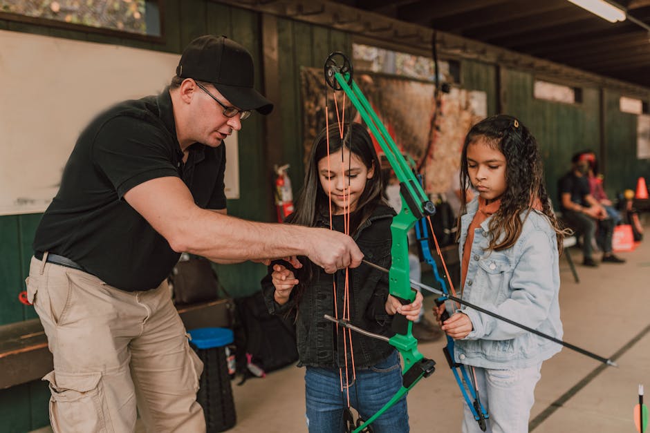 An instructor in a black cap and glasses demonstrates archery to two young girls using a green bow and arrows at an outdoor archery range.