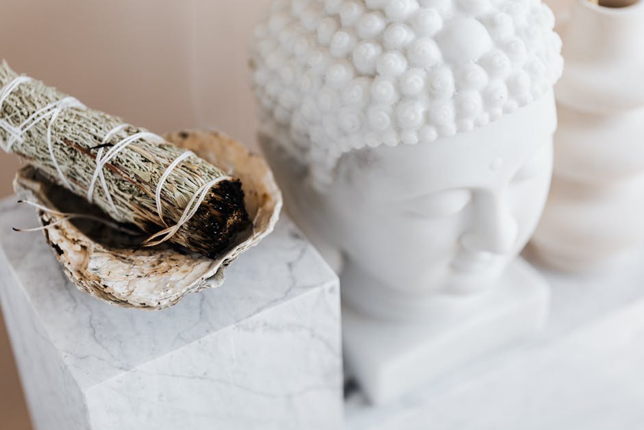 A close-up image of a decorative set featuring a sage smudge stick in an abalone shell, with a white bust of Buddha in the background, all placed on a marble surface.