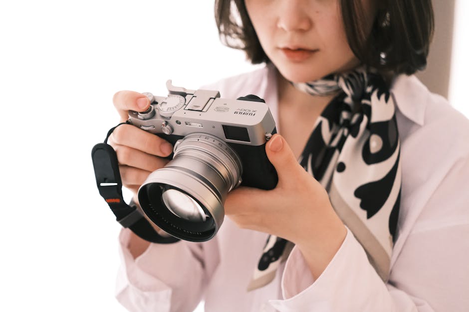 A person holding a silver and black mirrorless camera with a large lens, viewed in profile against a white background. They are wearing a white shirt with a black and white scarf, partially obscuring their face.