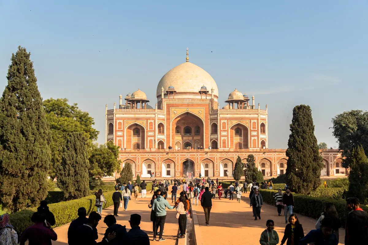 Visitors walking along a pathway leading to the historic red sandstone Mughal architecture of Humayun's Tomb under a clear blue sky.