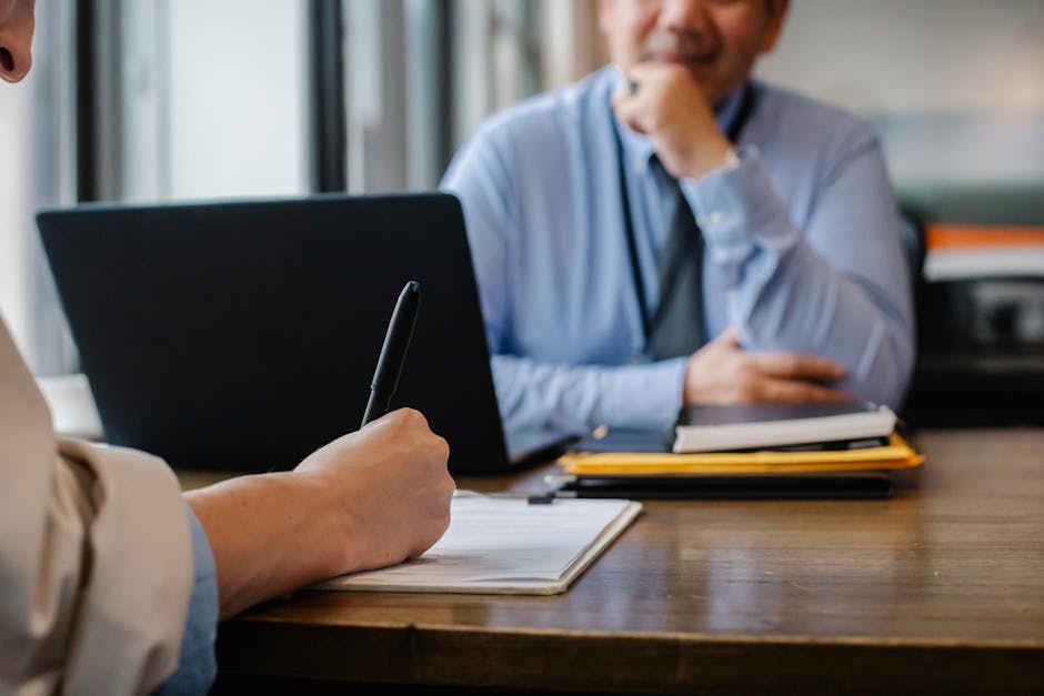 Person taking notes during a meeting with another person, who is smiling, sitting in the background with a laptop and documents on the table.