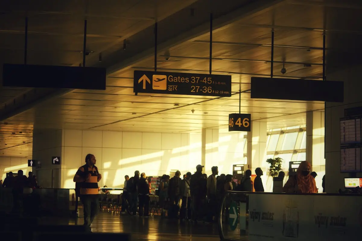 Airport terminal interior with passengers and silhouette figures near departure gates at sunset, direction signs for gates hanging from the ceiling, warm sunlight streaming through large windows.