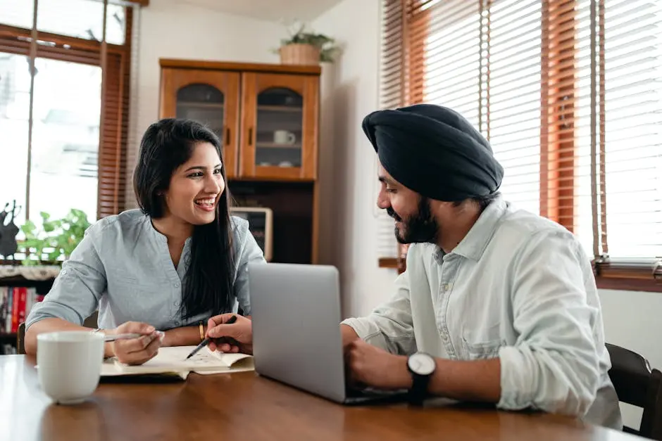 Two colleagues smiling and discussing work with a laptop and notebook at a home office table
